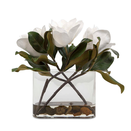 A Trio Of White Magnolia Blooms Are Placed In A Clear Glass Vase With Faux Water And Natural River Stones.